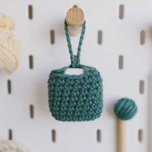 May include: A teal green, crocheted square pouch with a braided loop for hanging. The pouch is suspended from a wooden peg on a white pegboard. A ball of yarn and a wooden knitting tool are also visible.