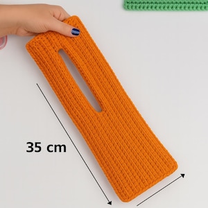 May include: An orange, rectangular, crocheted item with a central opening, measuring 35 cm (13.8 inches) in length. The handmade piece is held by a hand with blue nail polish. The item has a textured surface.