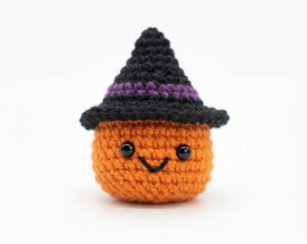 Puede incluir: Una pequeña figura de calabaza tejida a ganchillo de color naranja, con un sombrero de bruja negro puntiagudo y una banda morada. La calabaza tiene dos pequeños ojos de cuentas negras y una sencilla boca en forma de V bordada en negro.