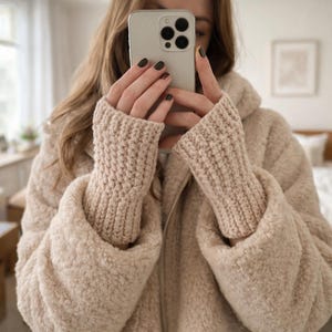 Peut inclure: Veste teddy bear beige avec des poignets en tricot assortis. La veste est zippée et a une texture douce et duveteuse. La personne tient un smartphone.