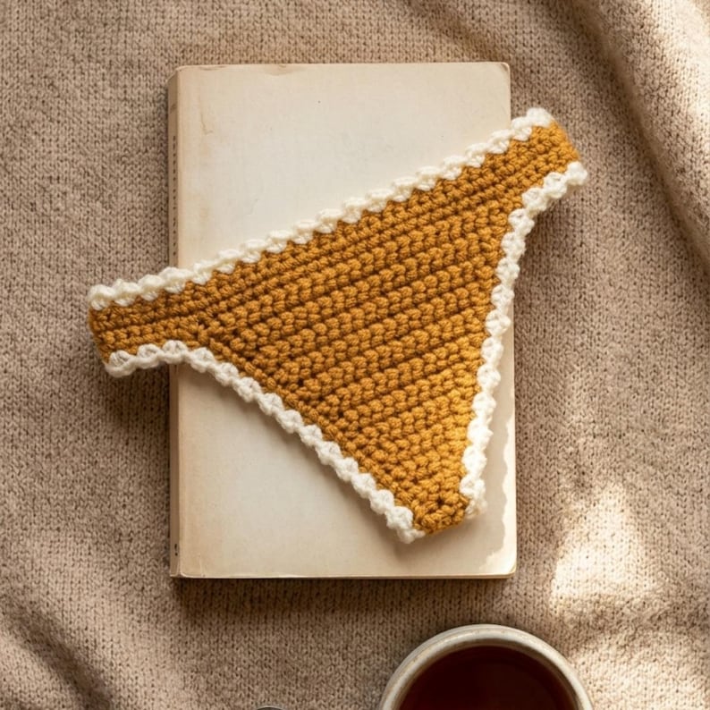May include: A mustard yellow crochet bikini bottom featuring a textured stitch pattern and a contrasting white scalloped trim along the edges. The garment is triangular in shape with a high-cut leg design and a simple, clean construction.