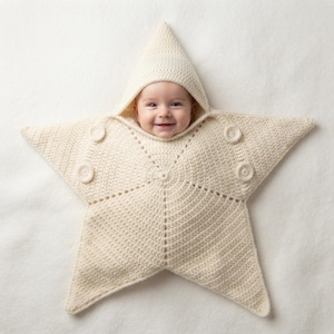 May include: A cream-colored, star-shaped crochet swaddle blanket featuring a pointed hood and four circular crochet buttons. The star-shaped garment is crafted from yarn with a visible stitch pattern, designed to wrap around an infant's body.
