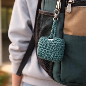 May include: A teal crochet case with a braided handle is attached to a backpack. The case is rectangular and appears to hold a small item. The backpack is green, tan, and black.