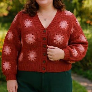 Könnte beinhalten: Ein rostfarbener V-Ausschnitt-Cardigan mit floralem Muster. Der Pullover hat quadratische Paneele, die jeweils mit einem hellrosa Blumendesign verziert sind. Der Cardigan hat eine Knopfleiste und lange Ärmel. Das Model trägt eine grüne Hose.