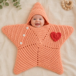 Peut inclure: Une gigoteuse pour bébé en forme d'étoile crochetée de couleur pêche avec une capuche, dotée de trois boutons blancs sur un côté et d'une petite application de cœur rouge crocheté sur le devant, avec un motif en maille texturée.