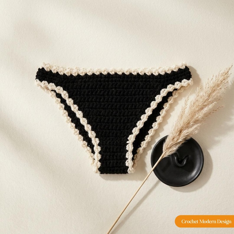 May include: Black crochet bikini bottom featuring a cream-coloured scalloped edge trim along the waistband and leg openings. The garment is displayed alongside a black ceramic dish and a dried pampas grass stem. Text reads Crochet Modern Design.
