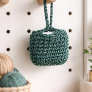 May include: A small, square, teal-colored crochet pouch with a braided handle, hanging from a wooden peg on a white pegboard. The pouch is made with a tight stitch, and the texture is visible. Other craft supplies are in the background.