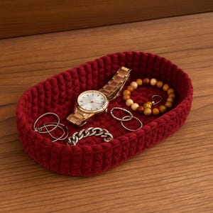 Peut inclure: Un plateau ovale rouge en crochet fait à la main contenant des bijoux et une montre-bracelet dorée. Les bijoux comprennent des bagues en argent, un bracelet chaîne en argent et un bracelet de perles en bois. La montre a un bracelet doré et un cadran blanc.