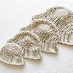 May include: Four beige crochet hats of varying sizes are arranged diagonally on a white surface. The hats have a textured, open weave design, with a slightly rounded crown and a small brim. The hats appear to be handmade.