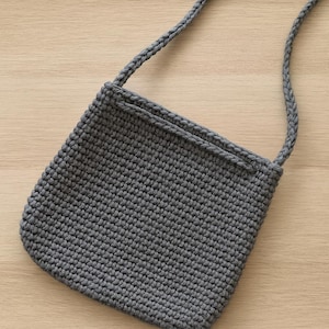 May include: A gray crocheted crossbody bag with a long, braided strap. The bag is square-shaped with rounded corners and a simple, textured stitch pattern. The strap is attached at the top corners of the bag.