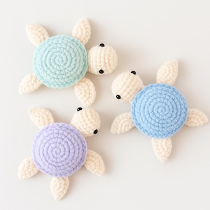 May include: Three handmade crochet turtle toys in pastel colors. Each turtle has a round shell in blue, light blue, and lavender, with off-white flippers and head. The turtles have black bead eyes and are arranged on a white background.