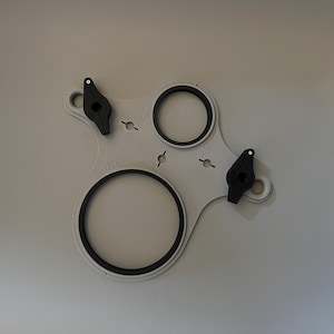May include: A light gray plastic tool with two black circular rings and two black levers. The tool has several small holes and measurements in millimeters. The rings have a black rubber lining. The tool is designed for a specific purpose.