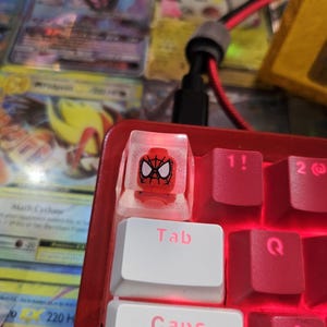 May include: A red and white keyboard keycap with a clear plastic casing featuring a cartoon image of Spider-Man. The keycap is labeled "Tab" and is surrounded by other red keyboard keys.