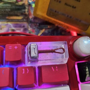 May include: A clear plastic keycap with a miniature silver and brown hammer inside. The keycap is sitting on a red mechanical keyboard with a white Enter key.