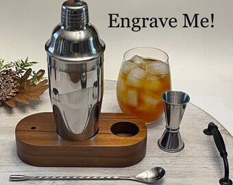 Könnte beinhalten: Ein Edelstahl-Cocktail-Shaker mit einem Holzständer, einem Jigger, einem Barlöffel und einem Glas mit Eistee. Der Shaker trägt die Aufschrift "Engrave Me!"