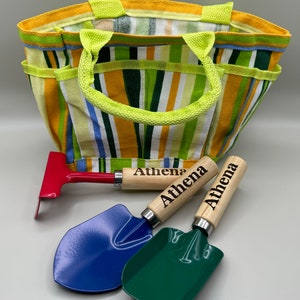 May include: A colourful striped canvas tote bag with a green handle. The bag contains three small gardening tools: a red rake, a blue shovel, and a green shovel. Each tool has a wooden handle with the name "Athena" engraved on it.