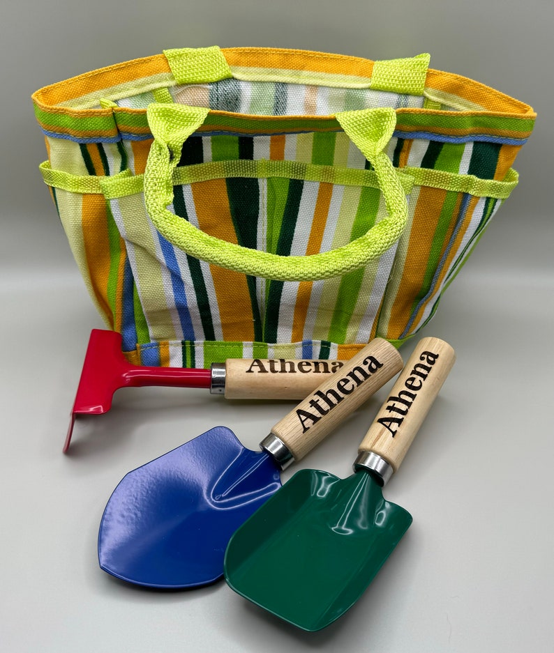 May include: A colourful striped canvas tote bag with a green handle. The bag contains three small gardening tools: a red rake, a blue shovel, and a green shovel. Each tool has a wooden handle with the name "Athena" engraved on it.