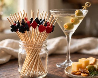 May include: A collection of wooden cocktail picks featuring red and black card suit symbols including hearts, diamonds, clubs, and spades. The picks are stored in a clear glass jar, with a martini glass containing olives nearby.