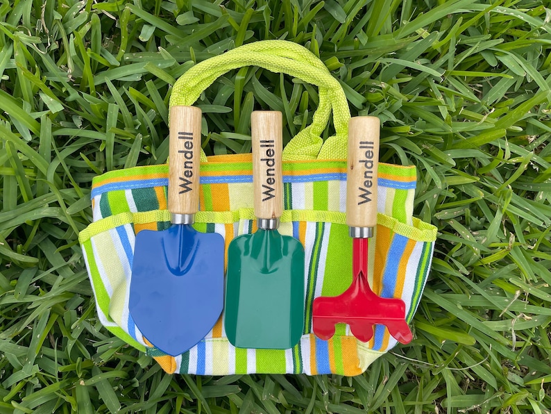 May include: A striped canvas gardening tote bag with three wooden handled gardening tools. The tools are a blue shovel, a green trowel, and a red rake. Each tool has the name "Wendell" engraved on the handle.