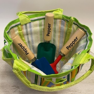 May include: Three small gardening tools with wooden handles in a green and white striped tote bag. The handles are labelled "Pepper".