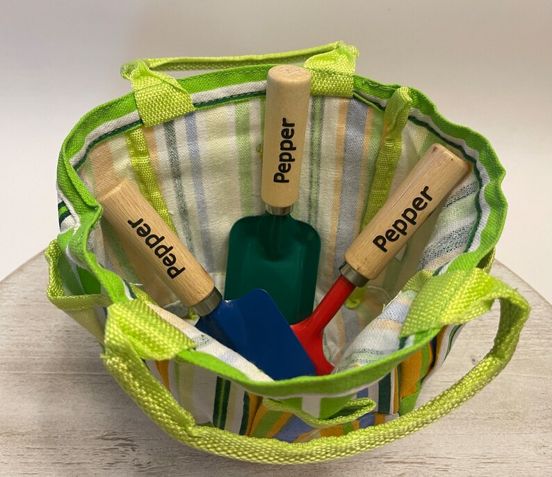 May include: Three small gardening tools with wooden handles in a green and white striped tote bag. The handles are labelled "Pepper".