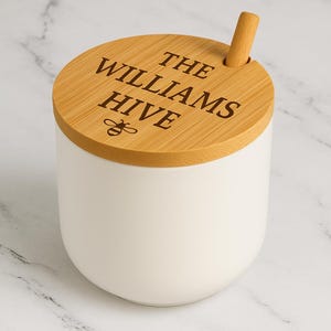 May include: A white ceramic honey pot with a wooden lid and a small wooden honey dipper. The lid is engraved with the words "THE WILLIAMS HIVE" and a bee graphic. The pot has a rounded shape and sits on a marble surface.