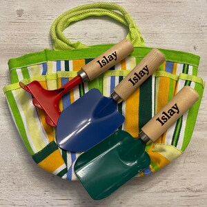 May include: A green and yellow striped canvas tote bag with three wooden handled gardening tools. The tools are a red rake, a blue trowel, and a green trowel. Each tool has the name "Islay" engraved on the handle.