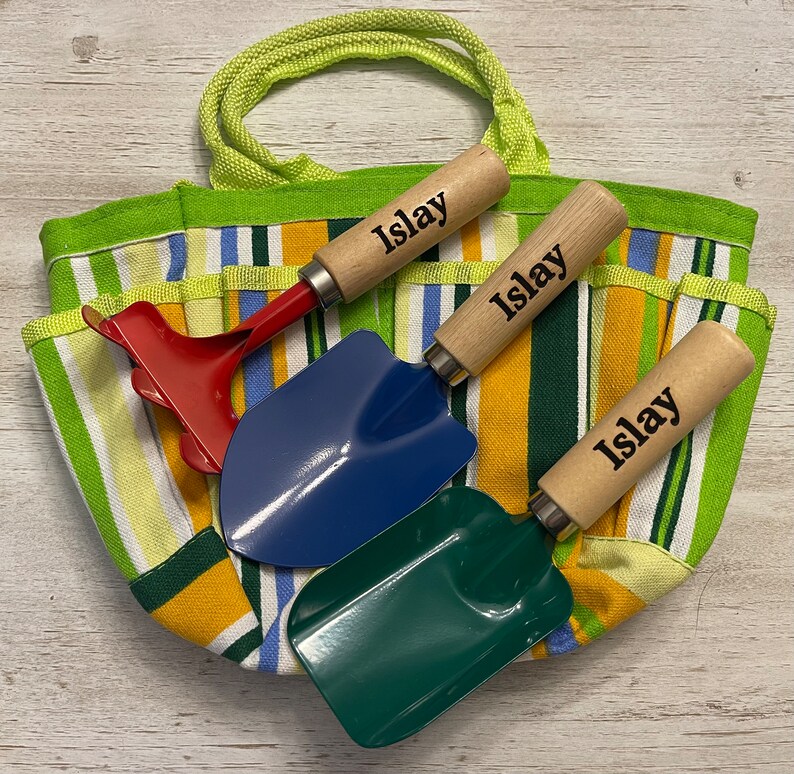 May include: A green and yellow striped canvas tote bag with three wooden handled gardening tools. The tools are a red rake, a blue trowel, and a green trowel. Each tool has the name "Islay" engraved on the handle.