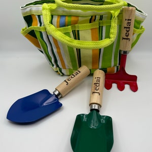 May include: A green and yellow striped canvas gardening tote bag with three gardening tools. The tools include a blue trowel, a green trowel, and a red rake. All three tools have wooden handles with the name "Jedai" engraved on them.