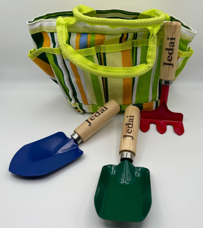 May include: A green and yellow striped canvas gardening tote bag with three gardening tools. The tools include a blue trowel, a green trowel, and a red rake. All three tools have wooden handles with the name "Jedai" engraved on them.