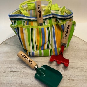 May include: A green and yellow striped canvas gardening tote bag with three gardening tools. The tools have wooden handles with the name "Charlotte" engraved on them. The tools include a trowel, a rake, and a hand cultivator.