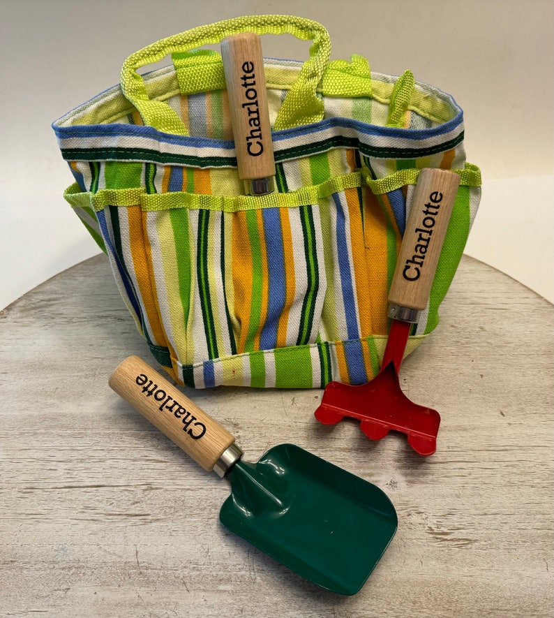May include: A green and yellow striped canvas gardening tote bag with three gardening tools. The tools have wooden handles with the name "Charlotte" engraved on them. The tools include a trowel, a rake, and a hand cultivator.