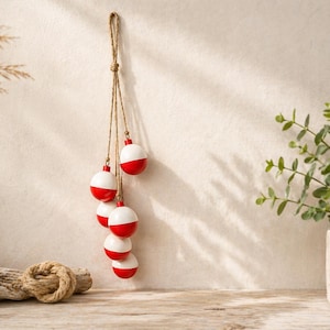 以下が含まれることがあります： 自然な麻ひもロープから吊り下げられた、赤と白の釣り用浮きが6つ付いた装飾用ハンギング。浮きはニュートラルな壁に対して均等に配置されています。貝殻、流木、鉢植えの植物が前景にあります。