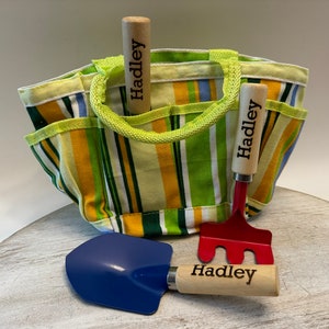 May include: A yellow and green striped canvas gardening tote bag with two wooden handled gardening tools. The tools are a red rake and a blue shovel. Both tools have the name "Hadley" engraved on the handles.