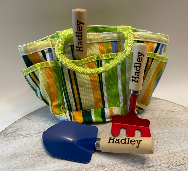 May include: A yellow and green striped canvas gardening tote bag with two wooden handled gardening tools. The tools are a red rake and a blue shovel. Both tools have the name "Hadley" engraved on the handles.