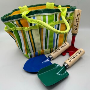 May include: A striped green, yellow and blue canvas gardening tote bag with a lime green handle. The tote bag is accompanied by three gardening tools with wooden handles that are personalised with the name "Sebastian". The tools include a red rake, a blue trowel and a green trowel.