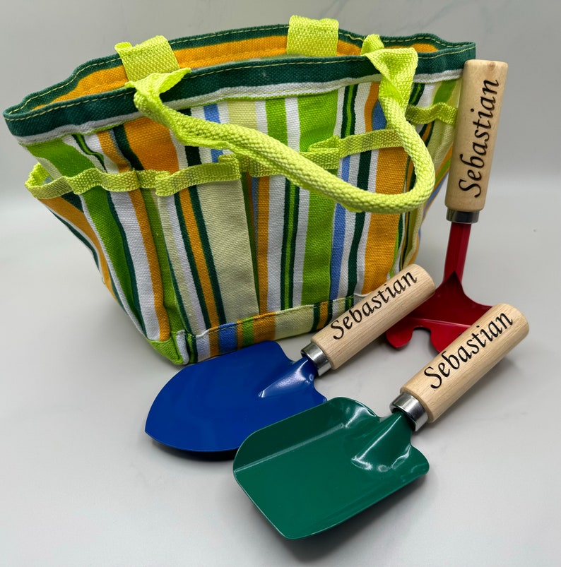 May include: A striped green, yellow and blue canvas gardening tote bag with a lime green handle. The tote bag is accompanied by three gardening tools with wooden handles that are personalised with the name "Sebastian". The tools include a red rake, a blue trowel and a green trowel.