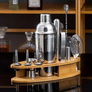 Op de afbeelding: Een roestvrijstalen cocktailshaker-set op een ovale bamboe standaard. De set bevat een driedelige shaker, een lange gedraaide menglepel, een Hawthorne-zeef, een ijstang, een dubbelzijdige jigger en drie flessenschenkers met stoppen.