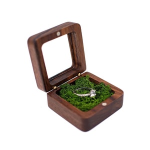May include: A square dark brown wooden ring box with a hinged lid and a clear window. The interior is filled with vibrant green preserved moss, cradling a silver-toned ring featuring a single clear stone and a band set with small sparkling accents.