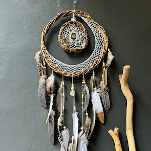 May include: A brown and white dreamcatcher with feathers hanging from the bottom. The dreamcatcher has a smaller circle in the center. The text "Mystic Handmade by Dream" is visible in the lower left corner of the image.
