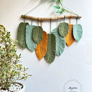 May include: A wall hanging made of macrame with a wooden branch and multiple green and orange feathers. The text "Mystic" is visible in the bottom right corner of the image.