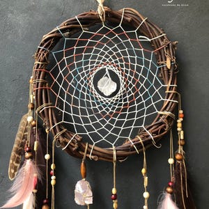 Puede incluir: Un atrapasue&ntilde;os de ramas marrones con hilo blanco, rojo y azul, un cristal blanco en el centro y plumas rosas, marrones y blancas que cuelgan de la parte inferior. El atrapasue&ntilde;os tiene cuentas de madera y un cristal rosa colgando.