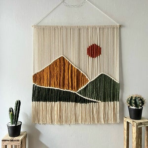 May include: A macrame wall hanging with a sunset and mountain scene. The mountains are made of yellow, green, and orange yarn. The sun is a red circle with orange rays. The wall hanging is made of natural cotton yarn and has a fringe at the bottom.