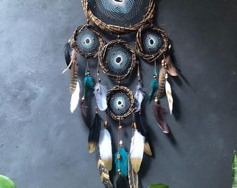 Puede incluir: Un gran atrapasueños con múltiples aros y plumas. Los aros son de madera marrón y las plumas son blancas, marrones y azul verdoso. El atrapasueños está colgado en una pared gris. El texto "Mystic Handmade by Wham" es visible en la esquina superior derecha de la imagen.