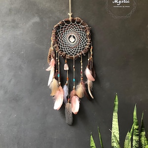 Puede incluir: Un atrapasue&ntilde;os marr&oacute;n con hilo blanco y rojo, plumas y cuentas que cuelgan de &eacute;l. El atrapasue&ntilde;os est&aacute; colgado de una cuerda contra una pared gris. El texto "Mystic Handmade by Nham" est&aacute; en la esquina superior derecha de la imagen.