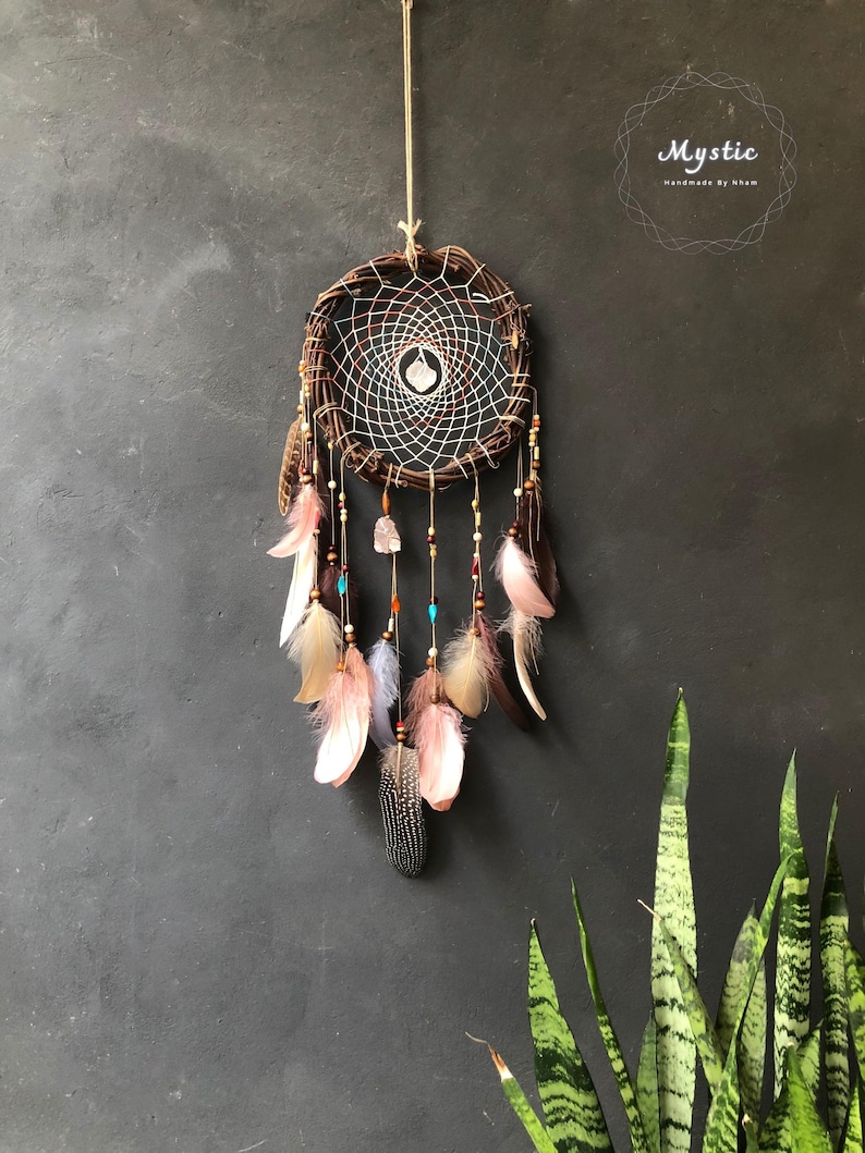 Puede incluir: Un atrapasue&ntilde;os marr&oacute;n con hilo blanco y rojo, plumas y cuentas que cuelgan de &eacute;l. El atrapasue&ntilde;os est&aacute; colgado de una cuerda contra una pared gris. El texto "Mystic Handmade by Nham" est&aacute; en la esquina superior derecha de la imagen.
