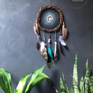 Puede incluir: Un atrapasueños marrón con red turquesa y blanca, plumas marrones y cuentas que cuelgan de la parte inferior. El atrapasueños está colgado en una pared gris con plantas verdes en primer plano. El texto "Mystic handmade by Nham" está en la esquina superior derecha de la imagen.
