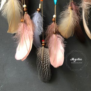 Puede incluir: Un atrapasue&ntilde;os con plumas en tonos marrones, rosas, blancos y azules. Las plumas est&aacute;n adornadas con cuentas y cuelgan de una base circular. El atrapasue&ntilde;os est&aacute; etiquetado como "Mystic Handmade by Nham".
