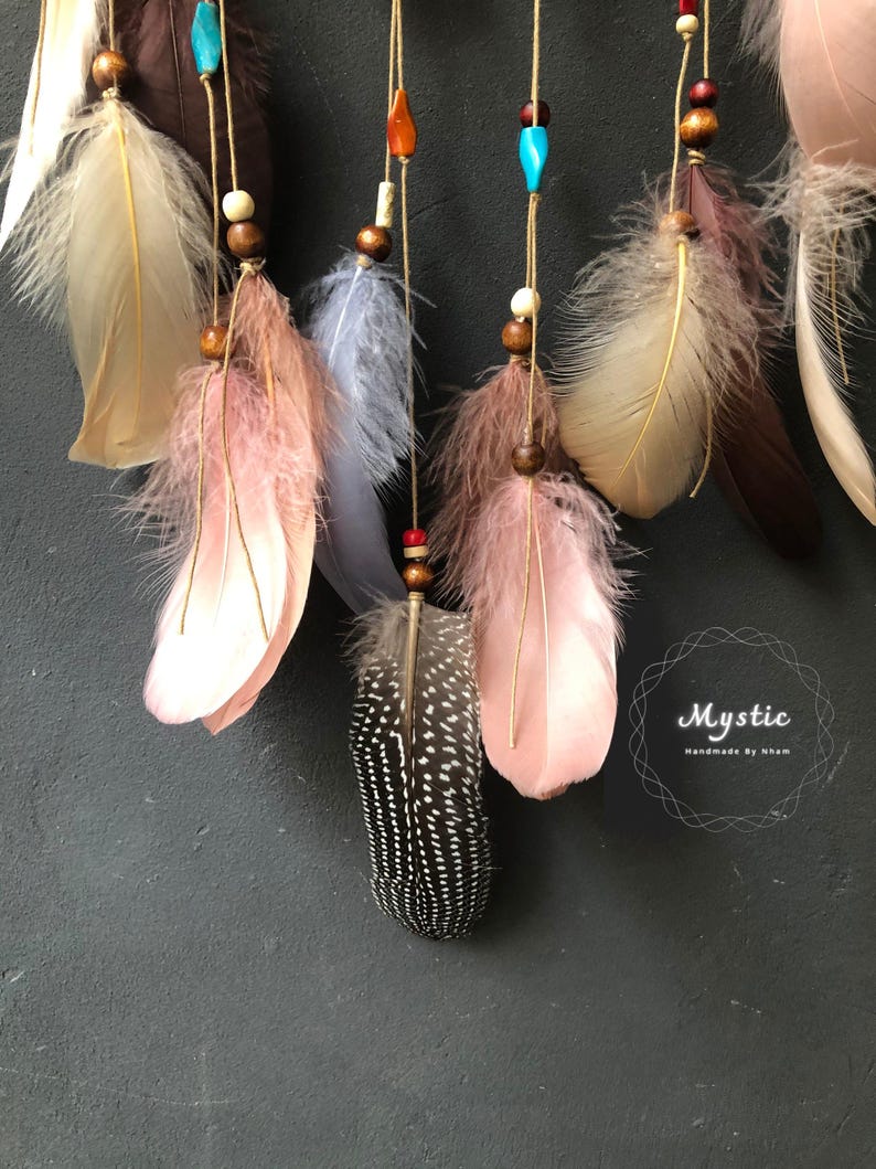 Puede incluir: Un atrapasue&ntilde;os con plumas en tonos marrones, rosas, blancos y azules. Las plumas est&aacute;n adornadas con cuentas y cuelgan de una base circular. El atrapasue&ntilde;os est&aacute; etiquetado como "Mystic Handmade by Nham".