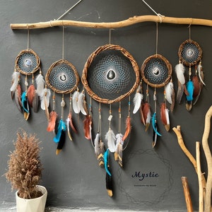 Puede incluir: Un conjunto de cinco atrapasueños con aros de madera marrón, redes blancas y azules y plumas en tonos marrones, blancos y turquesa. Los atrapasueños están colgados de una rama y el texto "Mystic Handmade by Dream" está en la parte inferior de la imagen.